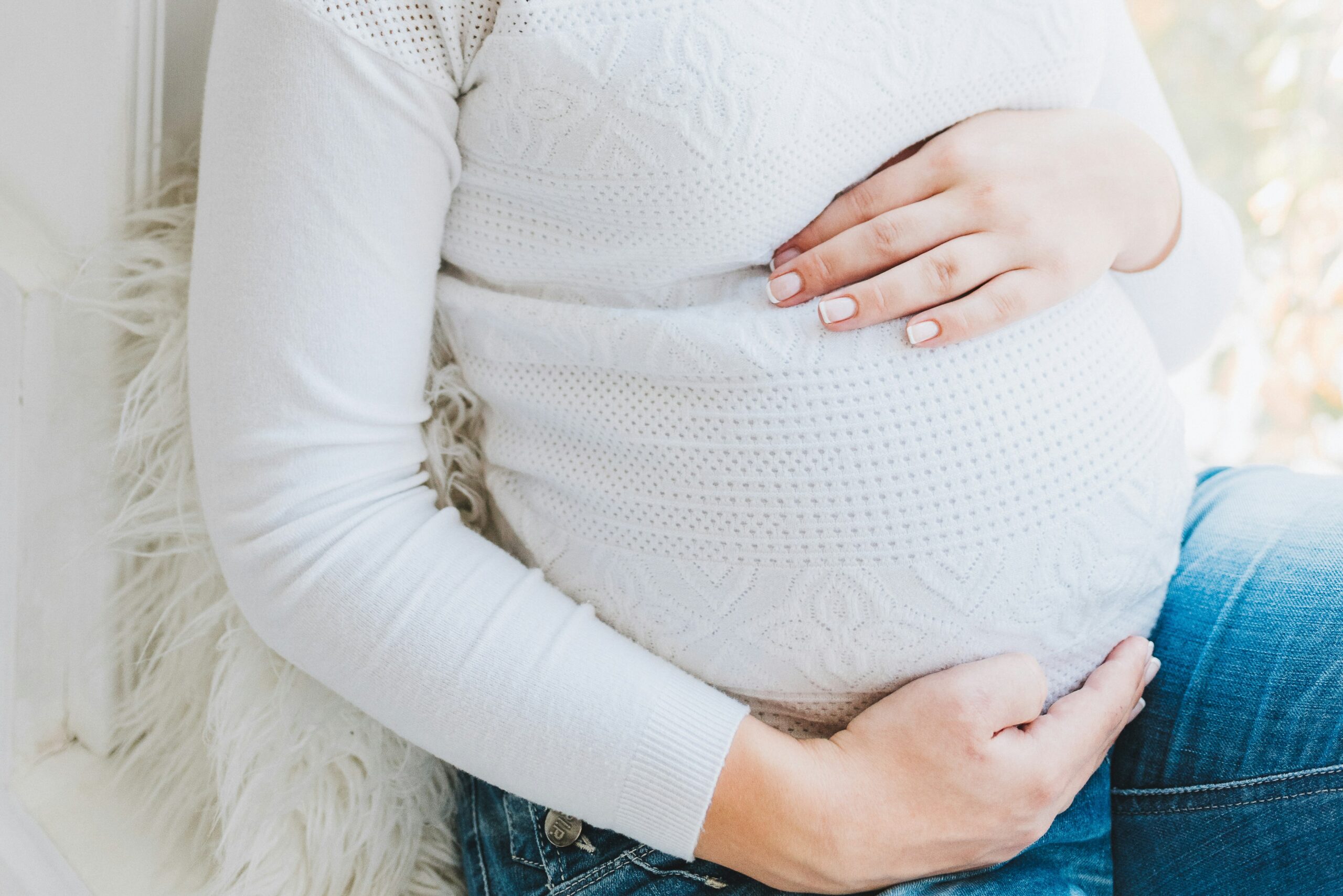 Mulher gestante sentada, com as duas mãos apoiadas sobre a barriga: uma na parte de cima e outra na parte de baixo, em um gesto carinhoso. Ela veste uma blusa longa branca que cobre a barriga e transmite suavidade. A imagem transmite acolhimento, serenidade e conexão com a gestação